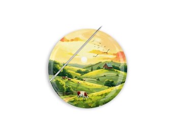 Può includere: Un piatto decorativo rotondo con una scena pastorale. L'immagine raffigura un tramonto su dolci colline verdi, un fienile rosso e mucche al pascolo. Un ago d'argento è posto diagonalmente sul piatto.
