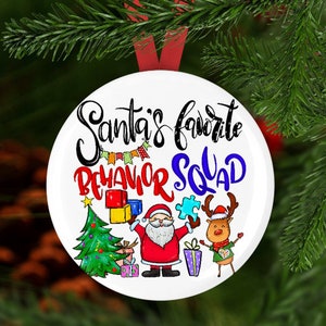 May include: White round Christmas ornament with the text "Santa's favorite behavior squad" in colorful lettering. The ornament features a cartoon Santa Claus, a reindeer, a Christmas tree, and colorful building blocks.