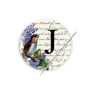 May include: A white button with a black letter J, a blue bird, purple flowers, and a handwritten script background. The button is attached to a needle.