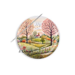 May include: A round needle minder with a watercolor painting of a small village nestled in a valley with trees and a church. The needle minder is decorated with a floral pattern and the text "J. Larson's".