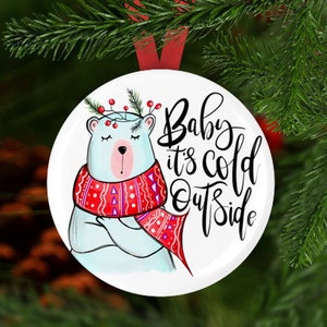 Może przedstawiać: Biała, okrągła ozdoba choinkowa z kreskówkowym niedźwiedziem polarnym w czerwono-białym wzorzystym szaliku. Tekst "Baby it's cold outside" jest napisany czarnym, ozdobnym pismem.