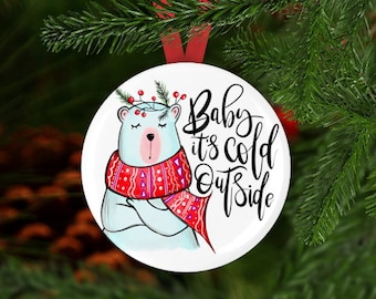 May include: White round Christmas bauble featuring a cartoon polar bear wearing a red and white patterned scarf. The text "Baby it's cold outside" is written in black cursive lettering.