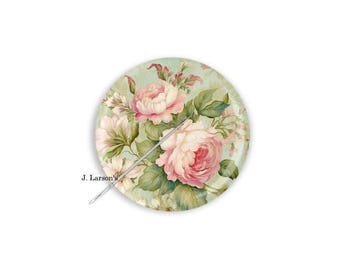 Può includere: Una spilla circolare con sfondo verde chiaro e un motivo floreale di rose rosa e foglie verdi. Un sottile ago argentato è posizionato diagonalmente al centro. Il testo J. Larson's è stampato in nero sul lato sinistro.