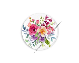 Può includere: Un cuscino per spilli in ceramica bianca con un design floreale ad acquerello con fiori rosa, gialli e viola. Un ago d'argento è posto sopra il cuscino per spilli.