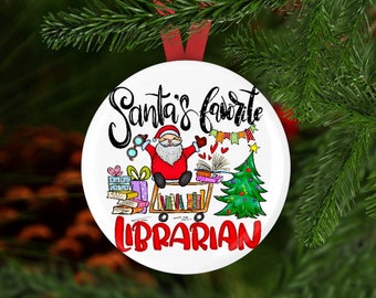 May include: White Christmas ornament with a red ribbon, featuring Santa Claus, books, and a Christmas tree. The text reads "Santa's favorite LIBRARIAN" in black and red. The ornament is hanging in front of a green Christmas tree.