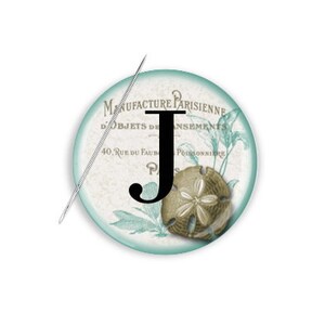 May include: A round, teal-colored button with a white background and a black letter J in the center. The button has a vintage design with text that reads "Manufacture Parisienne d'Objets de Fantaisie" and "40, Rue du Faubourg Poissonniere".