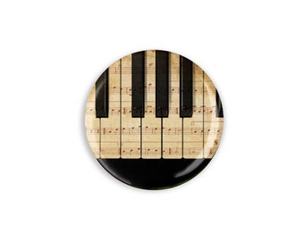 May include: A round button with a black and white piano keyboard design. The keys are on top of a sheet of music with notes printed on it.