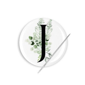 May include: A white round button with a black letter J surrounded by green watercolor leaves.