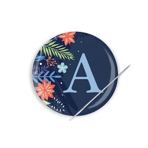 May include: A blue button with a floral design and the letter "A" in light blue. A silver needle is sticking through the button.