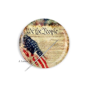 May include: Round decorative plate featuring the American flag and the text "We the People" in a vintage style. A silver needle is placed diagonally across the plate. The plate has a distressed, aged appearance.
