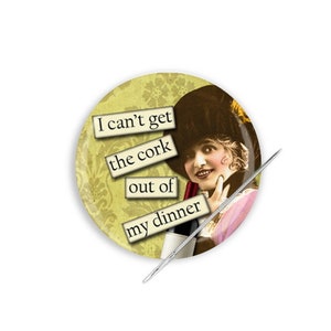 May include: A yellow button with a vintage illustration of a woman wearing a hat and holding a bottle of wine. The button has text that reads "I can't get the cork out of my dinner."