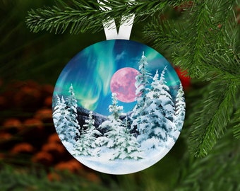 May include: Round Christmas ornament featuring a winter landscape with snow-covered pine trees, a pink moon, and the aurora borealis in shades of blue and green. The ornament has a white ribbon for hanging.