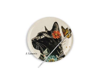 Può includere: Un bottone rotondo color crema con un profilo di gatto nero con un fiore rosso e una farfalla. Il bottone ha anche uno spillo d'argento e il testo "J. Larson's".