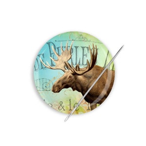 May include: A round button with a vintage-style image of a moose with antlers, surrounded by a blue and green background with the text "De Prairie & le" in a decorative font.