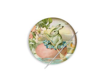 Può includere: Un bottone vintage a tema pasquale con un coniglio verde che rema una barca a forma di uovo rosa con fiori blu. Il bottone è circondato da un design floreale verde.
