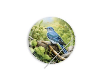 Può includere: Un uccello azzurro appollaiato su un ramo con foglie verdi. L'uccello ha un corpo blu chiaro e una coda blu più scura. Lo sfondo è un'immagine sfocata di una foresta.