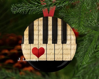 May include: Round Christmas ornament featuring a piano keyboard design with a red heart and text. The ornament has a black base and is suspended by a red ribbon against a backdrop of evergreen branches.
