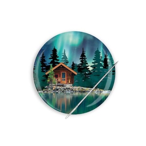 May include: A round needle minder with a cabin in the woods scene. The cabin is brown with a red roof and is surrounded by green trees. The background is a blue sky with green and purple aurora borealis.