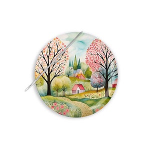 May include: A round needle minder with a watercolor illustration of a small village with houses, trees, and a winding path. The trees are in shades of pink, green, and yellow. The needle minder is white with a silver needle.