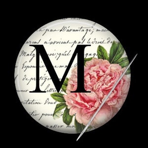May include: A white button with a black letter M and a pink flower with green leaves. The button has a needle sticking through it. The background is a white paper with black writing.