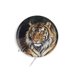 May include: A round, decorative button featuring a detailed painting of a tiger's face. The tiger has brown and black stripes, with yellow eyes and a white muzzle. The text "J. Larson's" is printed on the bottom left.