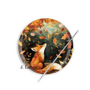 May include: A ceramic needle minder with a fox looking up at falling autumn leaves. The needle minder is round and has a dark green background with orange, yellow, and red leaves. The fox is orange and white. The text "J. Larson's" is printed on the bottom of the needle minder.
