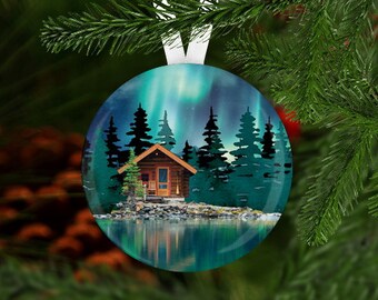 May include: A round Christmas ornament with a blue and green aurora borealis scene. A small wooden cabin with a lit window sits in the foreground. The ornament is perfect for a winter wonderland theme.