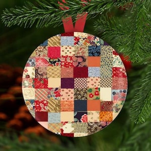 May include: A round Christmas ornament with a patchwork design. The ornament is made of various fabric squares in red, blue, orange, and cream colors. A red ribbon is attached for hanging. The ornament is displayed on a Christmas tree.
