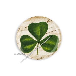 May include: A round, decorative shamrock pin with a green three-leaf clover design. The pin has a vintage, aged paper background with script writing. A silver needle is attached to the pin. The text "J. Larson's" is printed on the pin.