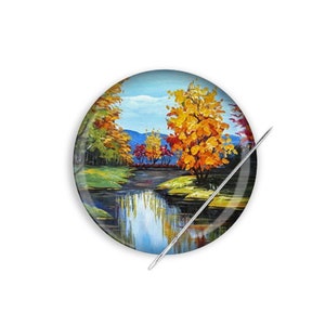 May include: A round pinback button with a painting of a fall landscape. The painting features a river, trees with yellow, orange, and red leaves, and a blue sky with white clouds.