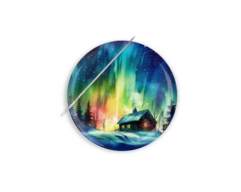 May include: A round button featuring a watercolour painting of a cabin in a snowy forest with a vibrant aurora borealis in the sky. The button is white with a silver needle.