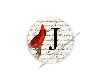 May include: A round pinback button featuring a red cardinal bird perched on a branch. The button has a white background with black script text and a black letter "J" in the centre.