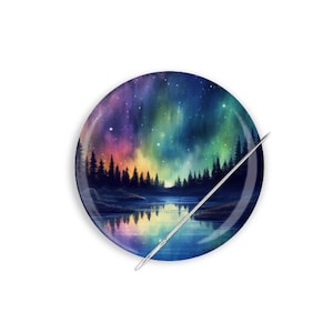 May include: A round needle minder with a colorful aurora borealis scene over a lake and forest. The needle minder is made of metal and has a shiny finish.