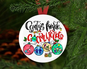 Può includere: Sfera natalizia rotonda bianca con il testo "Il lavoratore sociale preferito di Babbo Natale" in scrittura nera. La sfera è decorata con colorate illustrazioni di famiglie e un globo terrestre in stile cartone animato.