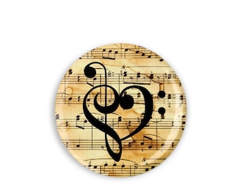 May include: Round decorative button with a musical theme. The button features a black treble clef and a heart shape against a background of vintage sheet music. The button has a glossy finish and a white background.