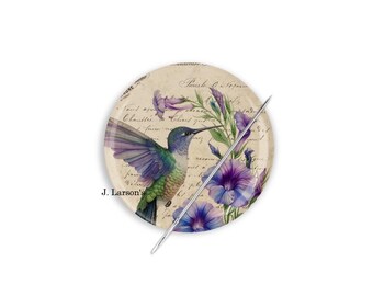 Può includere: Un piatto decorativo rotondo con un colibrì con piume viola e verdi, circondato da fiori viola e scritte su sfondo beige. Un ago d'argento è posto sul piatto. Il testo "J. Larson's" è visibile.