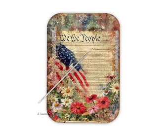 Può includere: Un oggetto decorativo rettangolare con un design della bandiera americana e la scritta "We the People". La bandiera è sovrapposta a uno sfondo simile a pergamena, circondata da fiori colorati. Un ago d'argento è posto diagonalmente sull'immagine.