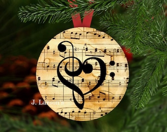May include: Round Christmas ornament featuring a musical score background with a black treble clef and bass clef forming a heart shape. The ornament is hanging from a red ribbon against a backdrop of green pine needles. The text "J. Larson's" is printed on the bottom left.