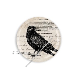 May include: A round, cream-colored pin with a black and white illustration of a raven on a background of cursive script. The text reads "INE CURSIVE DITE to helun bane, Cans Camm exte écrit en Expédite un geste brusqu". The pin is labeled "J. Larson's".