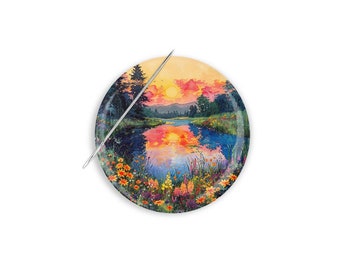 Può includere: Un magnete per aghi rotondo e decorativo con un paesaggio di tramonto su un lago, con un ago d'argento. L'immagine include fiori e alberi colorati.