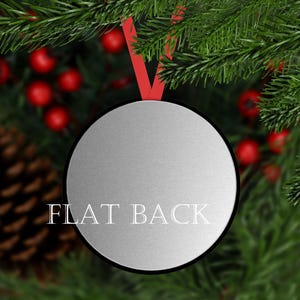 May include: A silver round bauble with a flat back. The bauble is hanging from a red ribbon against a background of green pine branches and red berries.