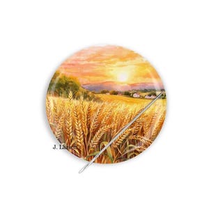 May include: A circular needle minder featuring a printed landscape of a golden wheat field under a sunset sky with orange and yellow hues. A silver metal needle is positioned diagonally across the surface. The text J. Larson's is printed in black.