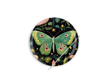 Può includere: Un cuscino per aghi rotondo con sfondo nero e una farfalla verde con accenti rosa. La farfalla è circondata da fiori e foglie. Un ago sporge dal cuscino per aghi.