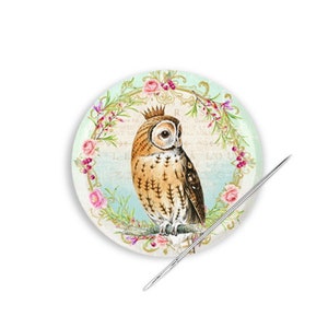 May include: A round pincushion with a floral design and an owl in the center. The owl is brown and white with large, yellow eyes. The pincushion is surrounded by a wreath of pink flowers and green leaves. The pincushion is on a white background.