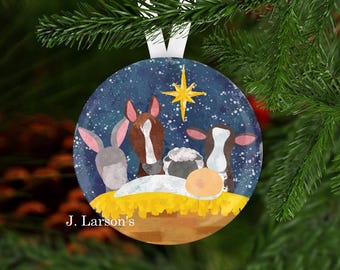 May include: Round Christmas ornament with a nativity scene. The ornament features a blue background with a star and a scene with a donkey, horse, sheep, and cow. The ornament has a white ribbon for hanging and the text "J. Larson's".