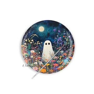 May include: A round needle minder featuring a colorful illustration of a white ghost standing in a vibrant field of wildflowers under a dark blue night sky with a full moon. The text J. Larson's is printed in black on the lower left side.