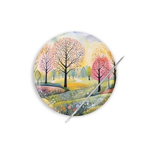 May include: A round needle minder with a watercolor painting of a whimsical forest scene. The needle minder features trees with pink, yellow, and green leaves, a winding path, and a field of flowers. A silver needle is placed on top of the needle minder.