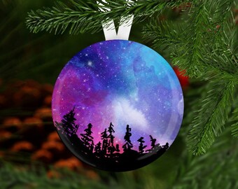 May include: A round glass ornament with a watercolor galaxy design in shades of purple, blue, and pink. Silhouettes of trees are visible in the foreground.