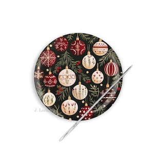 May include: A round, decorative Christmas ornament plate with a dark green background. The plate features various red and white ornaments with gold accents and green sprigs. A silver needle is placed diagonally across the plate.