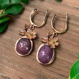 Puede incluir: Un par de pendientes colgantes con una piedra preciosa púrpura y detalles florales dorados.